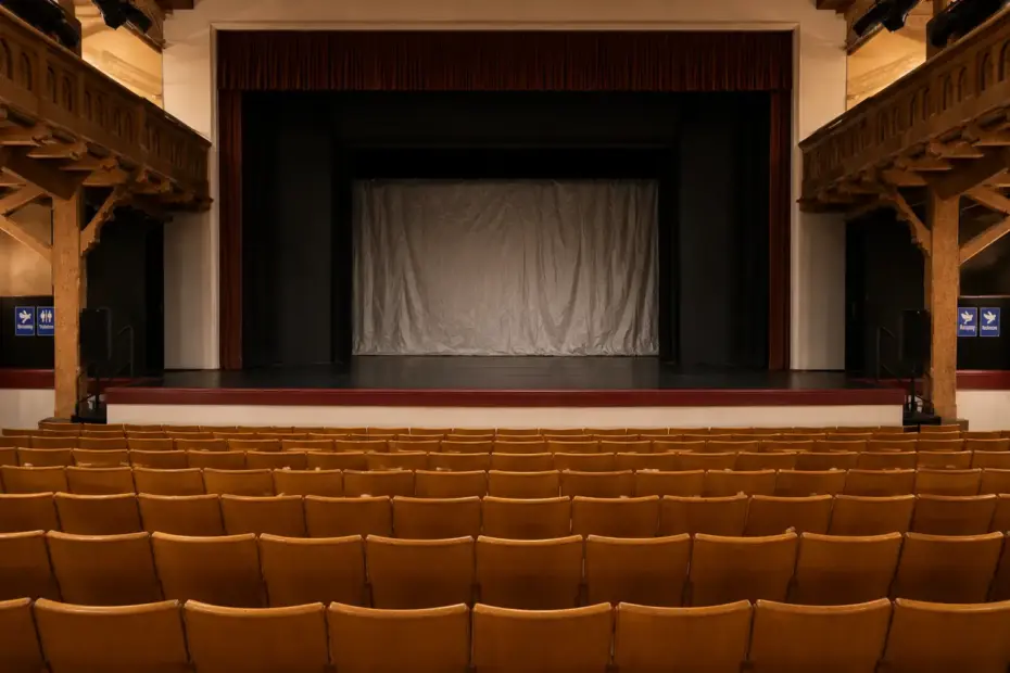 Ein leerer Theaterraum mit einer Bühne und braunen Sitzreihen. Die Bühne hat eine weiße Gardine.