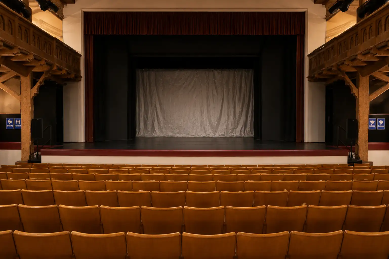 Ein leerer Theaterraum mit einer Bühne und braunen Sitzreihen. Die Bühne hat eine weiße Gardine.